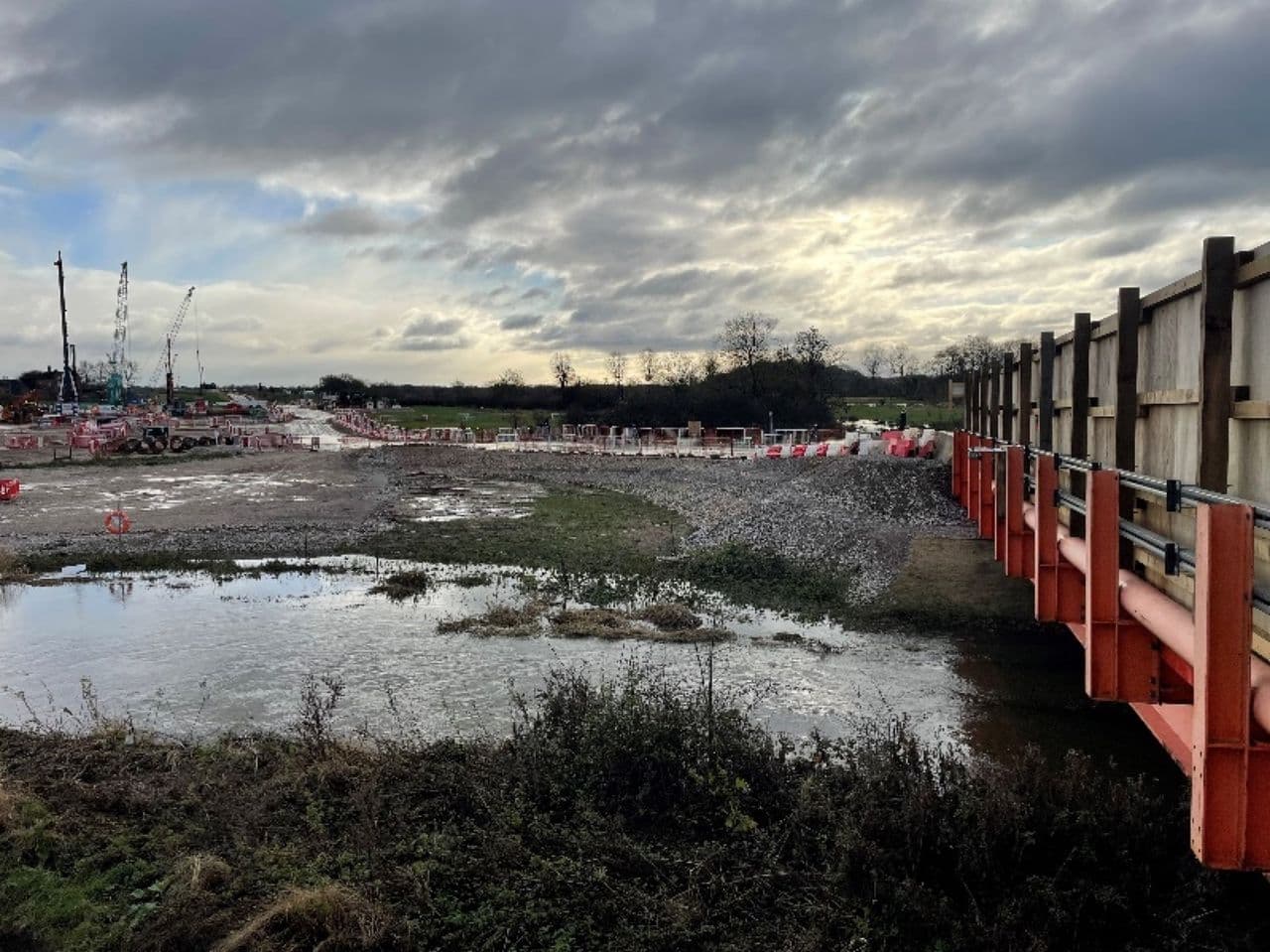 Standing floodwater on a construction site with cranes and site barriers under a cloudy sky