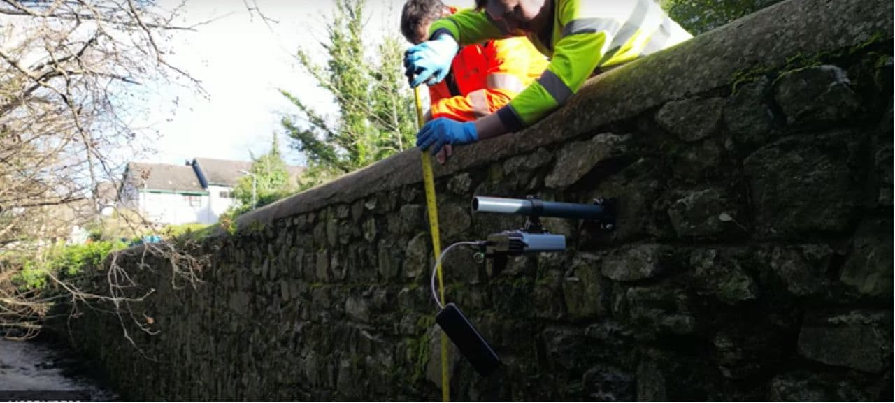 Workers in hi-vis jackets using a tape measure to install a sensor on a stone river wall