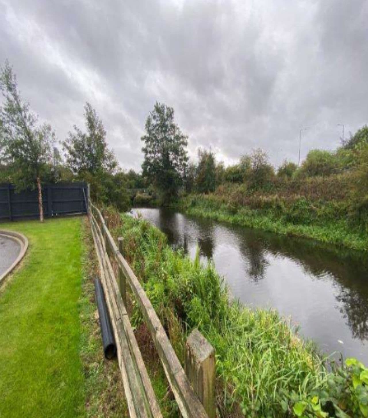 River lined with reeds beside a wooden fence and lawn under grey skies